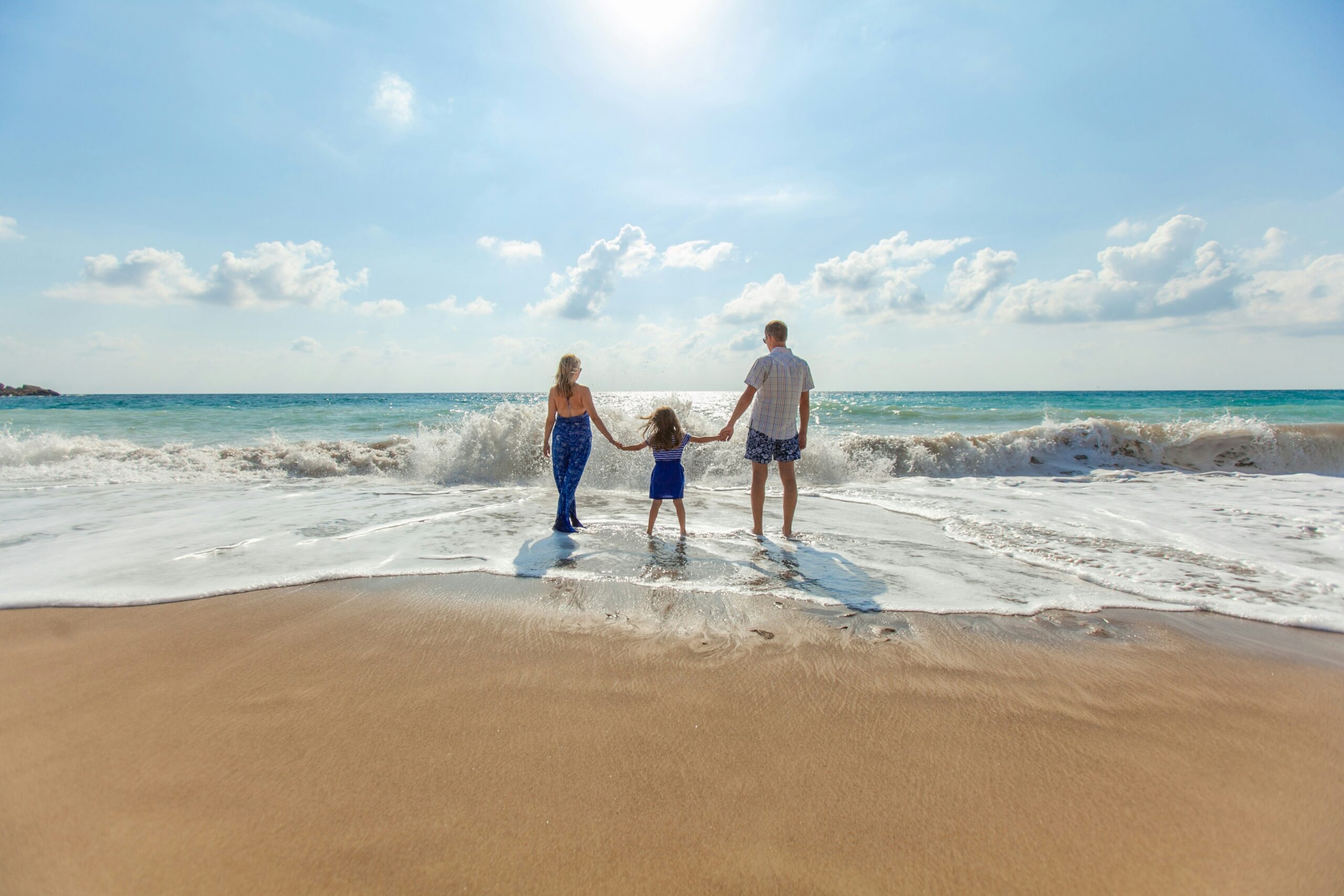 A family walking along a sandy beach at sunset, holding hands and smiling as waves gently approach the shore.